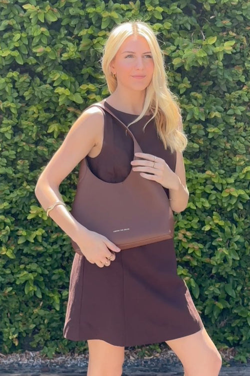 Woman wearing a brown sleeveless dress against a green hedge.