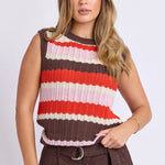 A woman modeling a sleeveless ribbed knit top with horizontal stripes in brown, red, orange, cream, and light pink, paired with brown shorts against a light gray studio background.