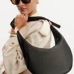 A model wearing a cream trench coat and black sunglasses carries a large, crescent-shaped black leather shoulder bag with gold-tone hardware against a plain white background.