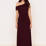 A woman modeling a floor-length off-the-shoulder dress in a deep burgundy or maroon shade, standing against a plain off-white wall on a light wood floor.