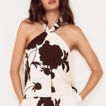A woman modeling a cream halter-neck two-piece set with a large dark brown floral print, featuring a sleeveless top and matching wide-leg pants, set against a plain white background.