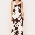 A woman modeling a sleeveless halter-neck maxi dress with a dark brown floral print on an off-white background, standing against a plain white wall on a light wood floor.