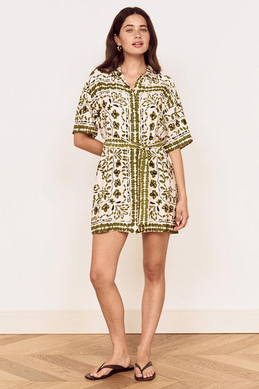 A woman modeling a short-sleeve button-front mini dress with an olive green and cream botanical pattern, standing on a light wood herringbone floor against a plain off-white wall.