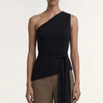 A model stands against a plain light background wearing a one-shoulder black wrap top and wide-leg taupe-brown pants with black sandals.