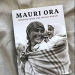 A book titled "MAURI ORA: WISDOM FROM THE MÁORI WORLD" lying on a textured beige fabric background. The cover features a black-and-white photograph of a smiling Máori woman carrying a child wrapped in a plaid blanket.