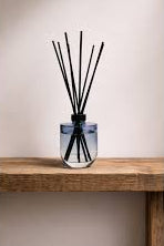 A reed diffuser with black reeds in a clear glass bottle with a blue tint, displayed on a rustic wooden shelf against a neutral beige wall.