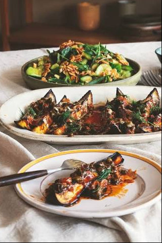 Three plates of prepared food on a tablecloth in a dining setting. The foreground plate holds a piece of grilled or roasted eggplant with sauce and herbs, the middle plate has a larger serving of the same dish, and the back plate shows a green salad with cucumber and fried onions.