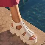 A person wearing chunky off-white platform sandals with a wide strap and buckle detail, standing on a stone poolside ledge with blue water in the background.