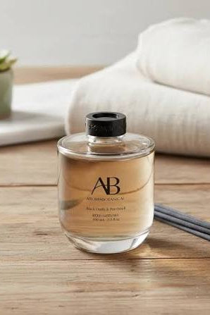 AB reed diffuser bottle filled with amber liquid, set on a wooden table with folded white towels and a small potted succulent in the background.