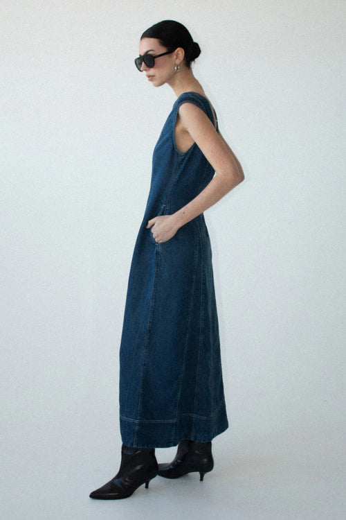 A woman stands in profile wearing a sleeveless dark blue denim maxi dress with black pointed-toe ankle boots and black sunglasses, photographed against a plain light gray studio background.