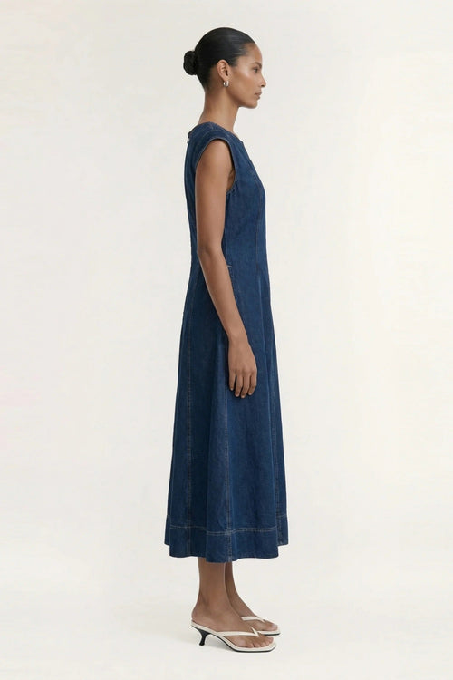 A woman in profile wearing a sleeveless dark blue denim midi dress and clear strap sandals, standing against a plain off-white studio background.