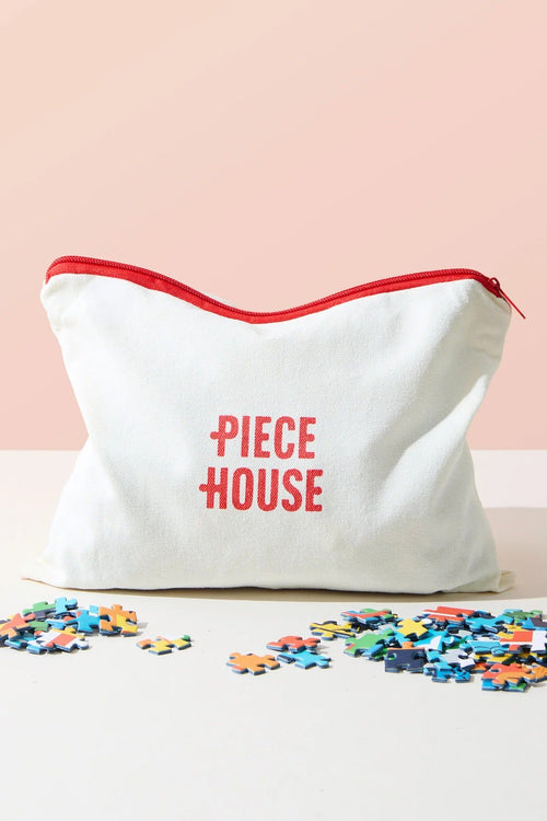 A white canvas zippered pouch with a red zipper and the words 'PIECE HOUSE' printed in red on the front, shown with scattered multicolored jigsaw puzzle pieces on a white surface against a soft peach-pink background.