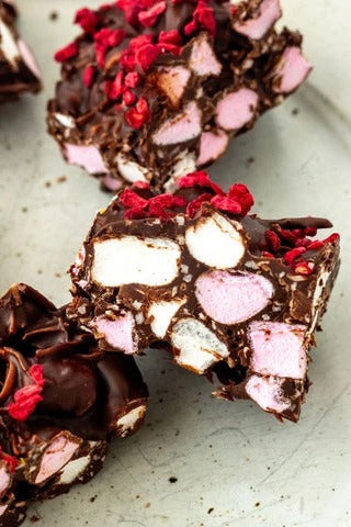 Close-up of several pieces of chocolate bark with pink marshmallows and bright red dried berry topping on a light gray surface.