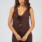 A woman modeling a sleeveless brown mini dress with a tie-front detail and decorative beads, photographed against a light gray studio background.