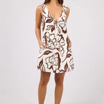 A woman modeling a sleeveless, knee-length dress with a brown floral print on a cream background, standing against a light gray studio backdrop.