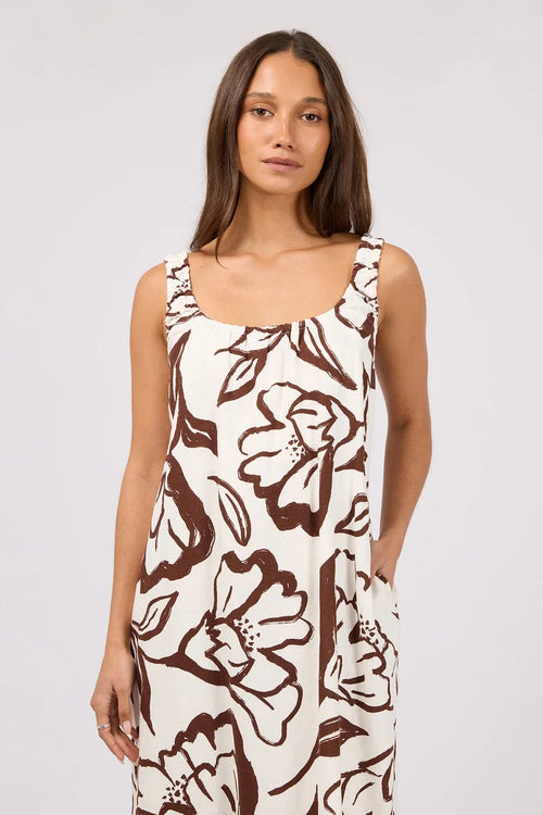 A woman modeling a sleeveless scoop-neck dress with a brown floral line-art print on an off-white background, photographed against a light gray studio backdrop.