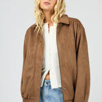 Woman wearing a brown jacket, white shirt, and blue jeans on a light gray background