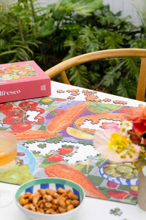 Jigsaw puzzle on a table with a box labeled 'Alfresco', flowers, and drinks in an outdoor setting.