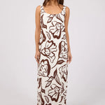 A woman modeling a sleeveless maxi dress with a brown floral and leaf pattern on an off-white background, paired with brown strappy sandals, against a light gray studio backdrop.