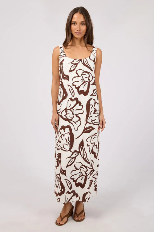 A woman modeling a sleeveless maxi dress with a brown floral and leaf pattern on an off-white background, paired with brown strappy sandals, against a light gray studio backdrop.