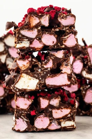 A close-up stack of chocolate-covered marshmallow treats with pink and white marshmallows and red dried fruit pieces, photographed on a light neutral surface.