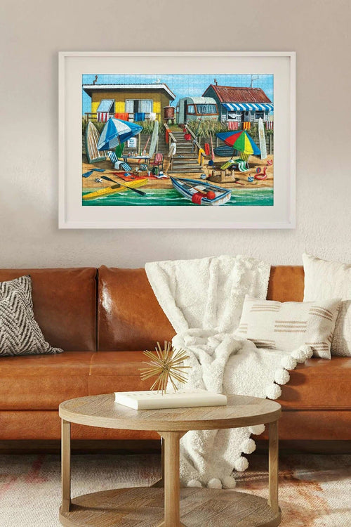 A colorful framed art print depicting a vibrant beach house scene, displayed on a beige wall above a brown leather sofa in a modern living room setting.