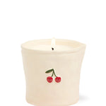 An off-white ceramic candle holder with a small cherry decoration on the front, containing white wax and a lit flame, set against a plain white background.