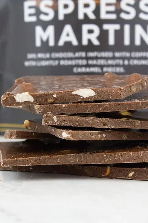 A stack of dark chocolate bark pieces with visible nuts and caramel pieces, set in front of a black bag labeled 'ESPRESSO MARTINI' on a light marble surface.