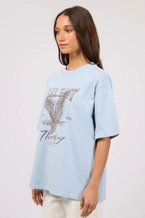 A woman wearing an oversized light blue graphic t-shirt with a distressed eagle design and the text 'SILENT THEORY' on the front, paired with off-white pants, set against a plain light gray studio background.