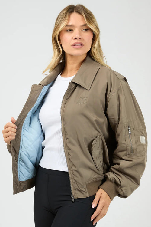 Woman wearing a brown bomber jacket with a white shirt and black pants on a light gray background