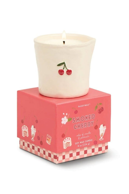 A lit cream-colored wax candle with a cherry illustration sits on top of its pink packaging box, which is labeled 'Smoked Cherry' and features a pink and white checkered base.