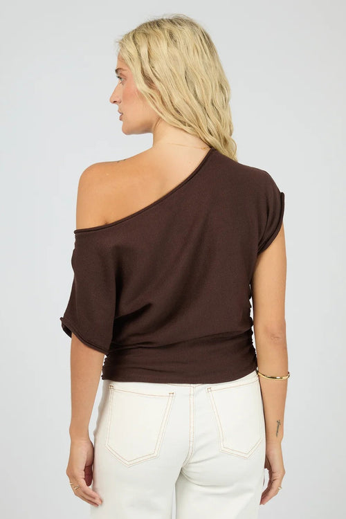 Woman wearing a brown one-shoulder top and white pants on a plain background