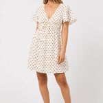 A woman modeling a short-sleeve cream dress with a black polka dot pattern, featuring a V-neckline, ruffled sleeves, and a tie detail at the waist, set against a plain light gray studio background.