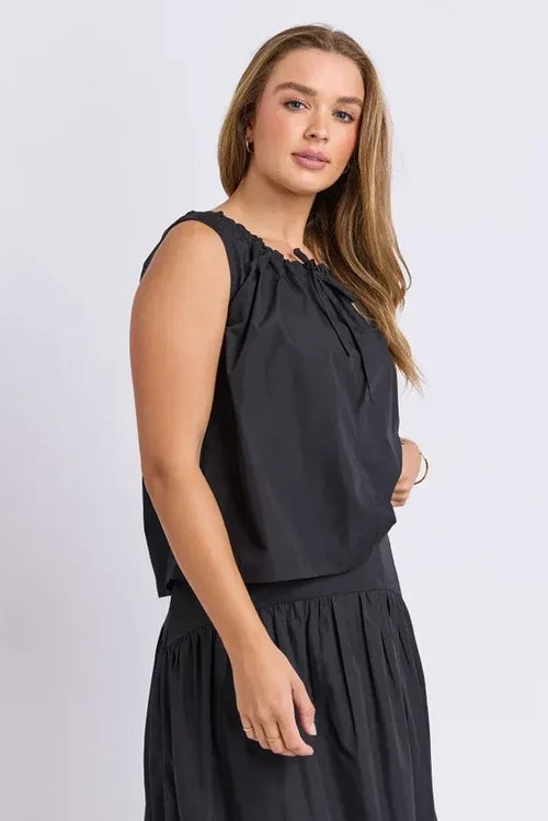 A woman modeling a sleeveless black top with a gathered, ruffled neckline and a matching black pleated skirt, photographed against a light gray studio background.
