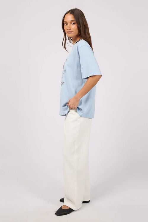 A woman modeling a light blue oversized short-sleeve t-shirt and white wide-leg trousers, standing against a plain light gray studio background.