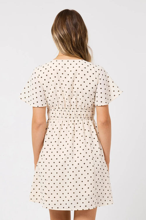 A woman viewed from the back wearing a short-sleeve cream dress with a black polka dot pattern and a smocked waist, set against a plain light gray background.