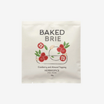 A single packet of Baked Brie Cranberry and Almond Topping, Herb & Spice, shown on a clean white background. The packaging is a cream-colored stand-up pouch with a graphic of a brie cheese wheel topped with red cranberries and green leaves.