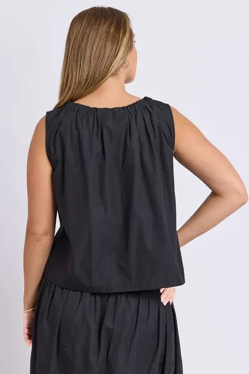 A woman shown from the back wearing a sleeveless black top with a gathered, pleated neckline and a matching black skirt, set against a plain light gray studio background.
