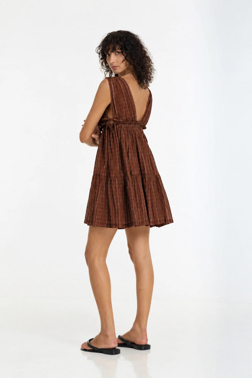 A woman modeling a brown pleated halter mini dress with a tie waist, shown from the back against a plain white studio background.