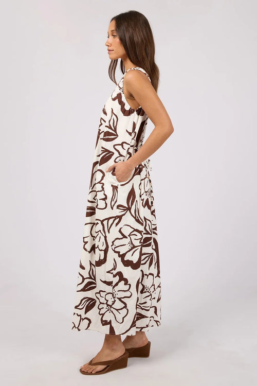 A woman modeling a sleeveless maxi dress with a brown floral pattern on an off-white background, shown from a side profile against a light gray studio backdrop.