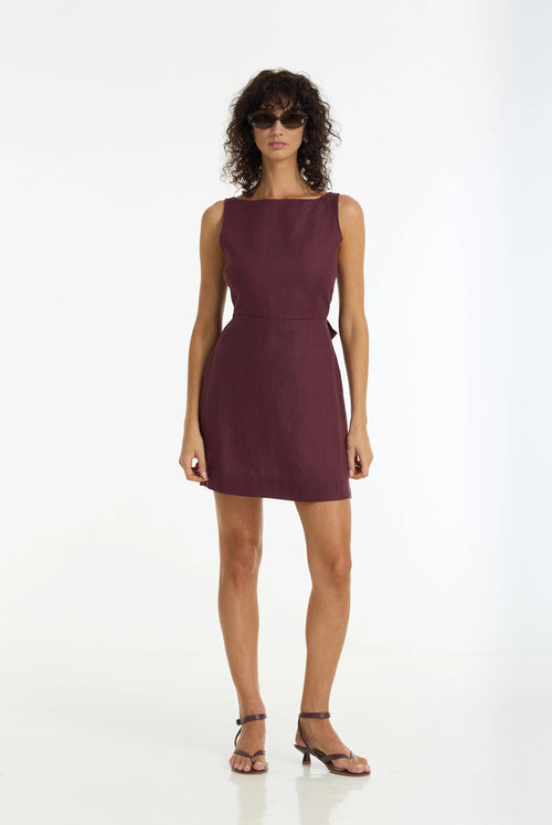 A woman modeling a sleeveless, knee-length wine-colored dress with a structured neckline and a matching belt, standing against a plain white studio background.