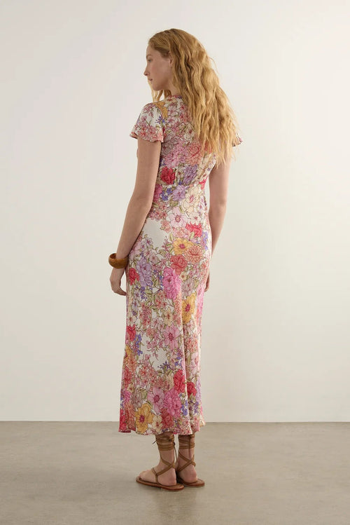 A woman stands in profile wearing a short-sleeve maxi dress with a dense multicolor floral print on a light cream base. The dress features pink, red, yellow, and blue flowers.