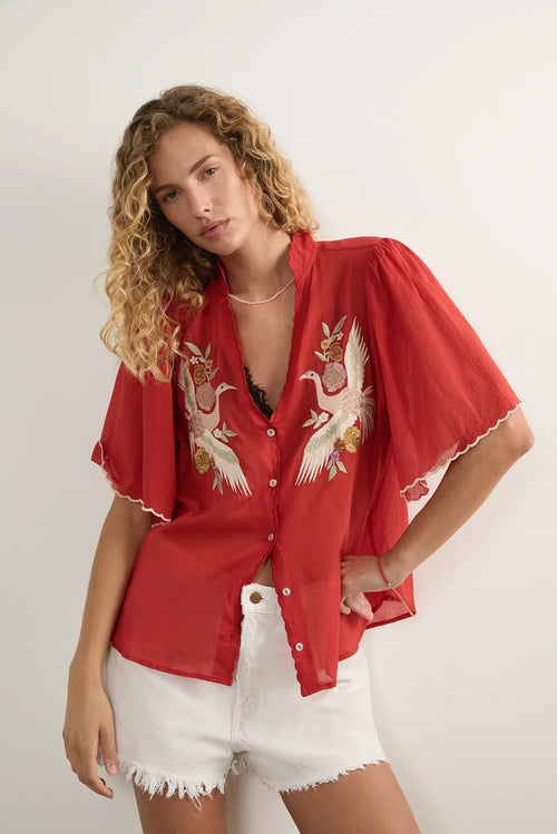 A woman modeling a red short-sleeve button-up shirt with white and gold crane embroidery on the front, paired with white denim shorts against a plain light background.