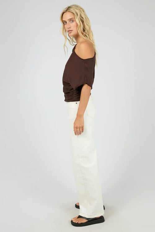 A woman modeling a dark brown off-the-shoulder wrap top and white wide-leg pants, standing against a light gray studio background.