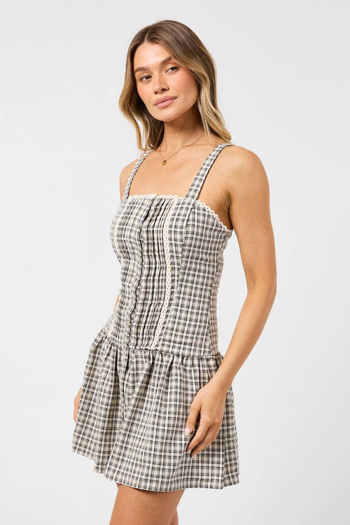 A woman modeling a sleeveless mini dress with a brown and white gingham plaid pattern, featuring a smocked bodice and ruffled straps, set against a plain light gray studio background.