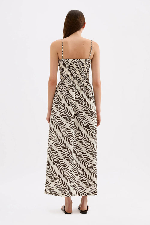 A woman viewed from the back wearing a sleeveless maxi dress with thin straps and an allover abstract black-on-cream wavy pattern, standing against a plain light gray studio background.