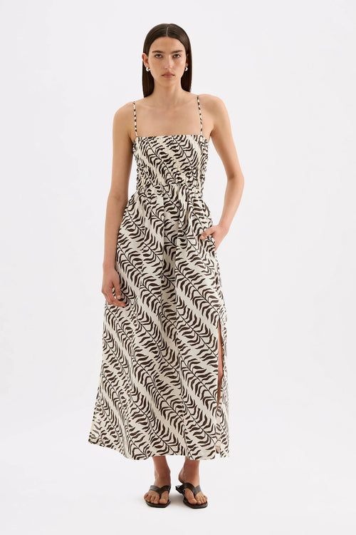 A woman modeling a sleeveless maxi dress with a black and off-white abstract wavy pattern, thin shoulder straps, and a side slit, photographed against a plain light gray studio background.