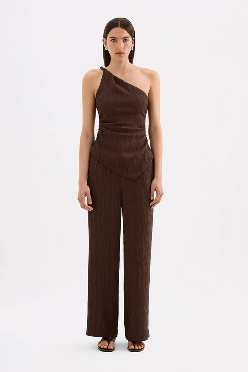 A woman modeling a dark brown one-shoulder wrap-style jumpsuit with matching wide-leg pants, photographed against a clean white studio background.