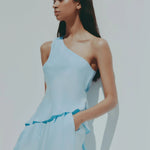 A woman modeling a light blue one-shoulder jumpsuit with a matching fabric belt, set against a plain light gray background.