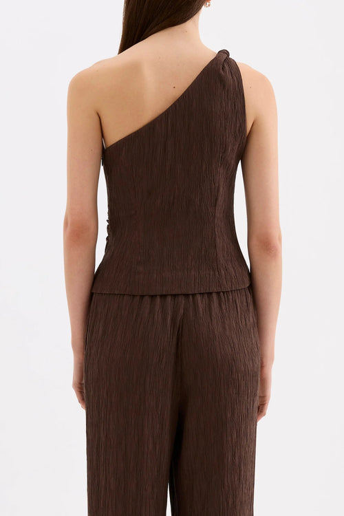 A model shown from the back wearing a dark brown one-shoulder jumpsuit with a textured, crinkled fabric, set against a plain light gray background.